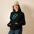 Cargar la imagen en la vista de la galería, Smiling person in black unisex eco hoodie with teal graphic, beige beanie, and blue jeans