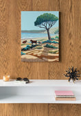 Charger l'image dans la visionneuse de la galerie, Vertical canvas print of serene coastal landscape with tree and cows, hanging on wood-paneled wall above modern white shelf